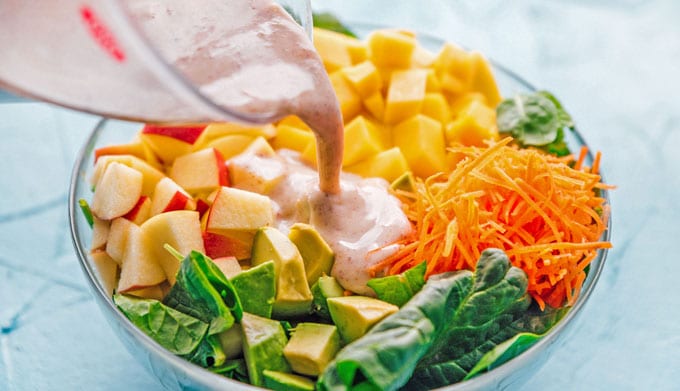 Pouring dressing into a salad with spinach, mango, carrots, and apple in a bowl on a blue background