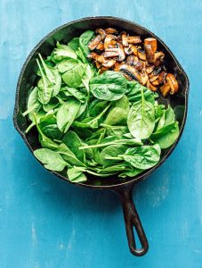 A cast iron skillet on a blue surface features fresh spinach on one side, while sautéed mushrooms complement a serving of spaghetti squash on the other.