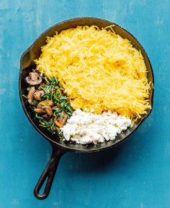 A cast iron skillet brimming with spaghetti squash and mushrooms delicately sautéed alongside vibrant greens and crumbled cheese, all set against a striking blue background.