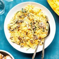A plate of creamy spaghetti squash with mushrooms and spinach, accompanied by a fork and spoon, set on a blue surface. Nearby are empty glasses and a bowl of uncooked pasta.