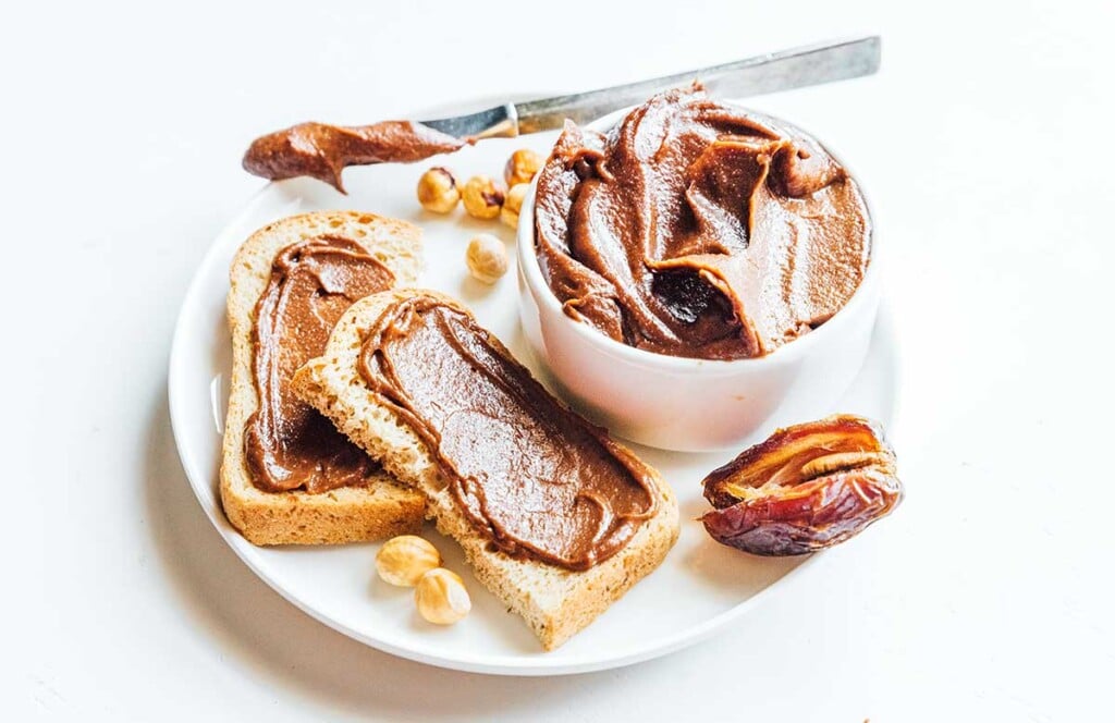 Two halves of bread coated with vegan hazelnut spread in addition to a small dish filled with the spread, a date, a few hazelnuts, and a knife, all atop a white plate