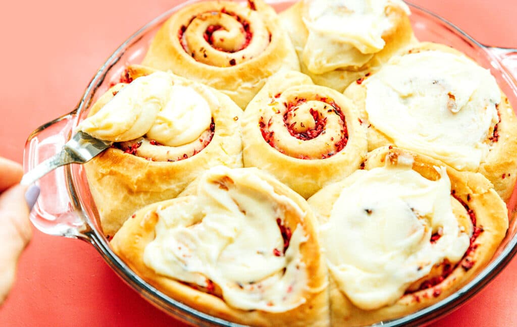 A hand icing a cinnamon roll placed in a baking dish filled with seven cranberry rolls