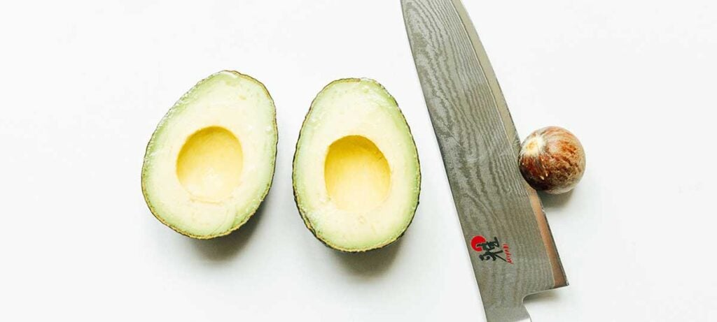 Two avocado halves facing inside-up next to a knife lodged to an avocado pit
