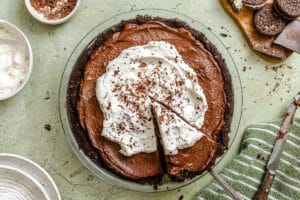 A chocolate cream pie with whipped cream and chocolate shavings sits in a glass pie dish. A slice is being lifted out with a pie server, revealing its rich avocado chocolate filling. Nearby are Oreo cookies, a bowl of whipped cream, and a striped towel.