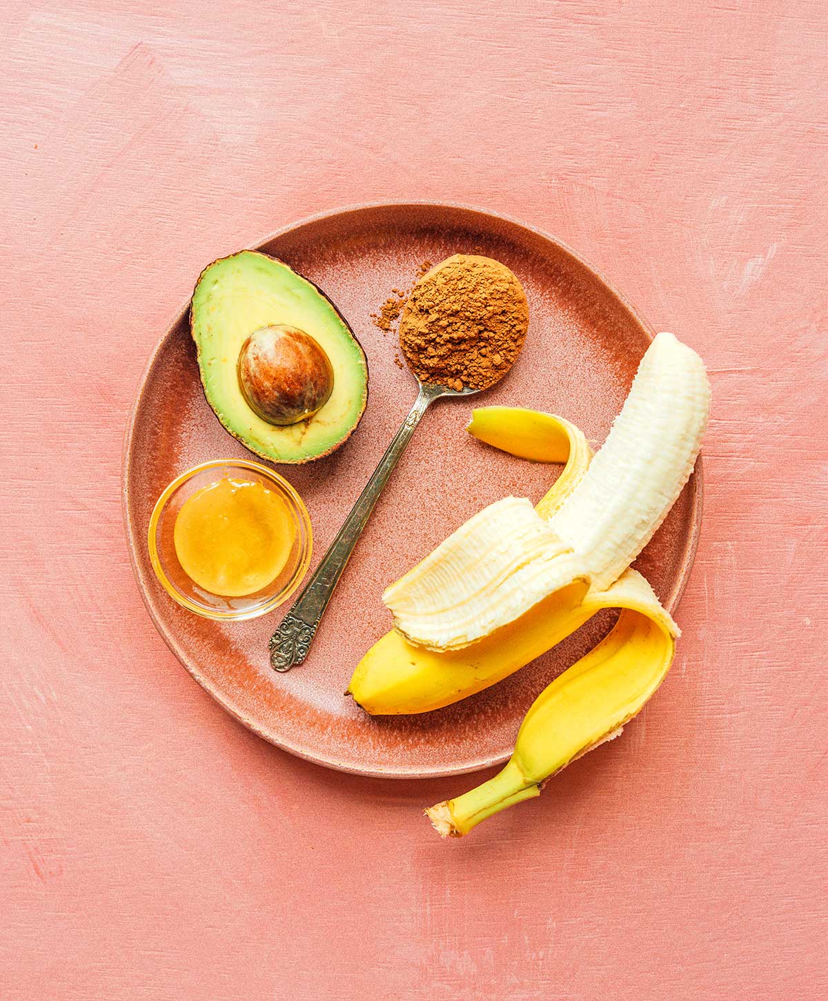 Honey, half of an avocado, a tablespoon of cocoa powder, and a peeled banana on a plate
