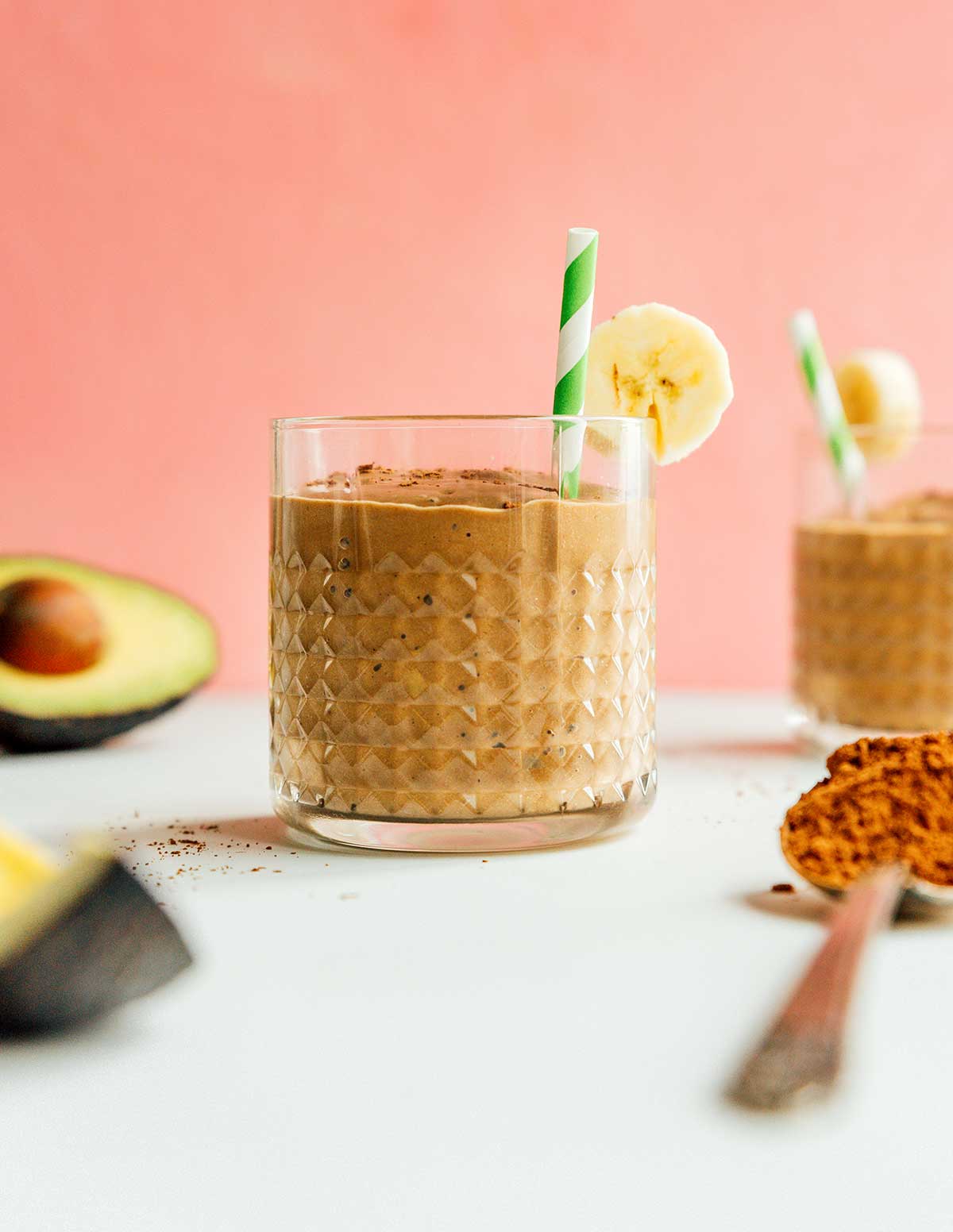 A glass filled with chocolate avocado smoothie and topped with a banana slice and a green and white striped straw