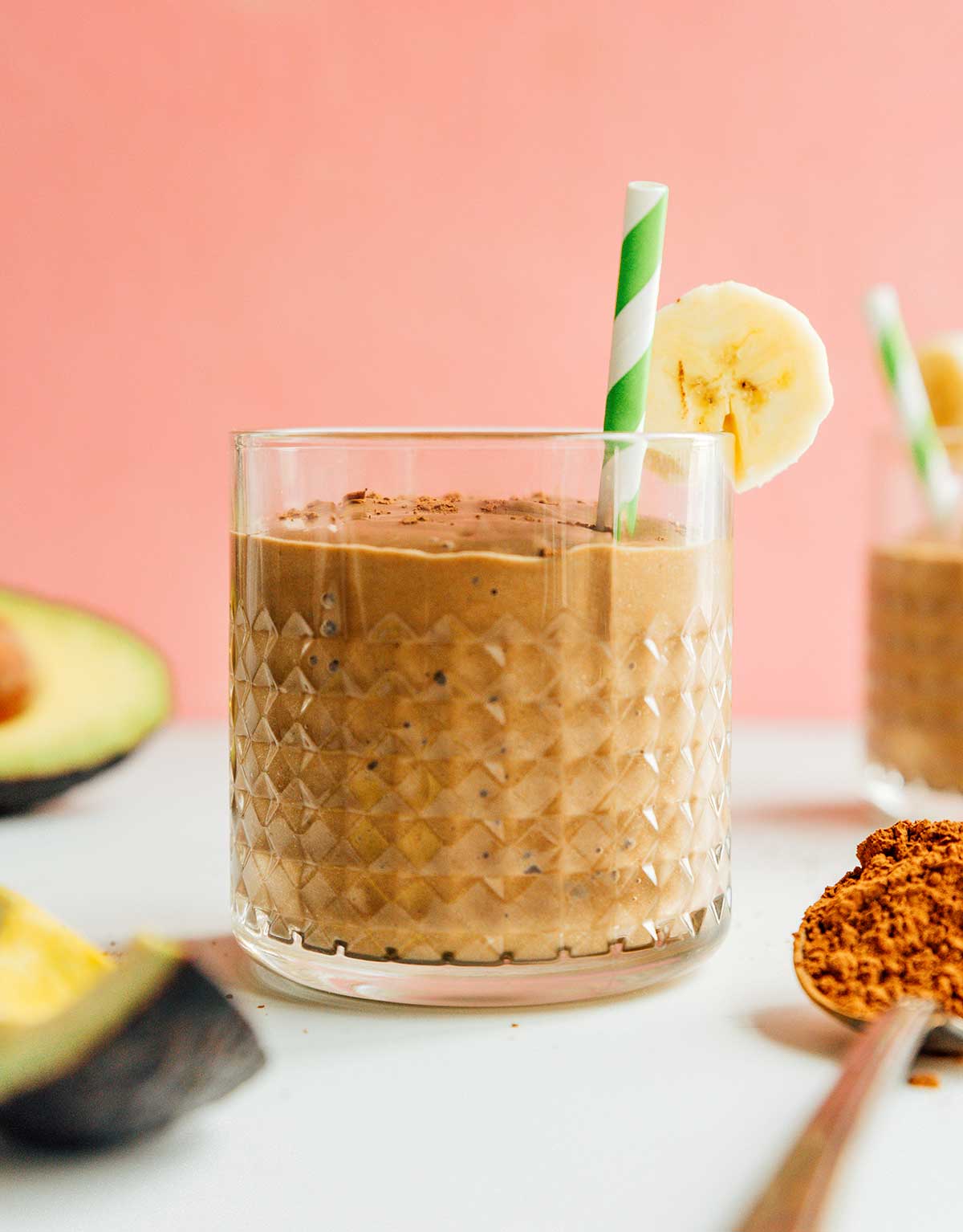 A glass filled with chocolate avocado smoothie and topped with a banana slice, cinnamon, and a green and white striped straw