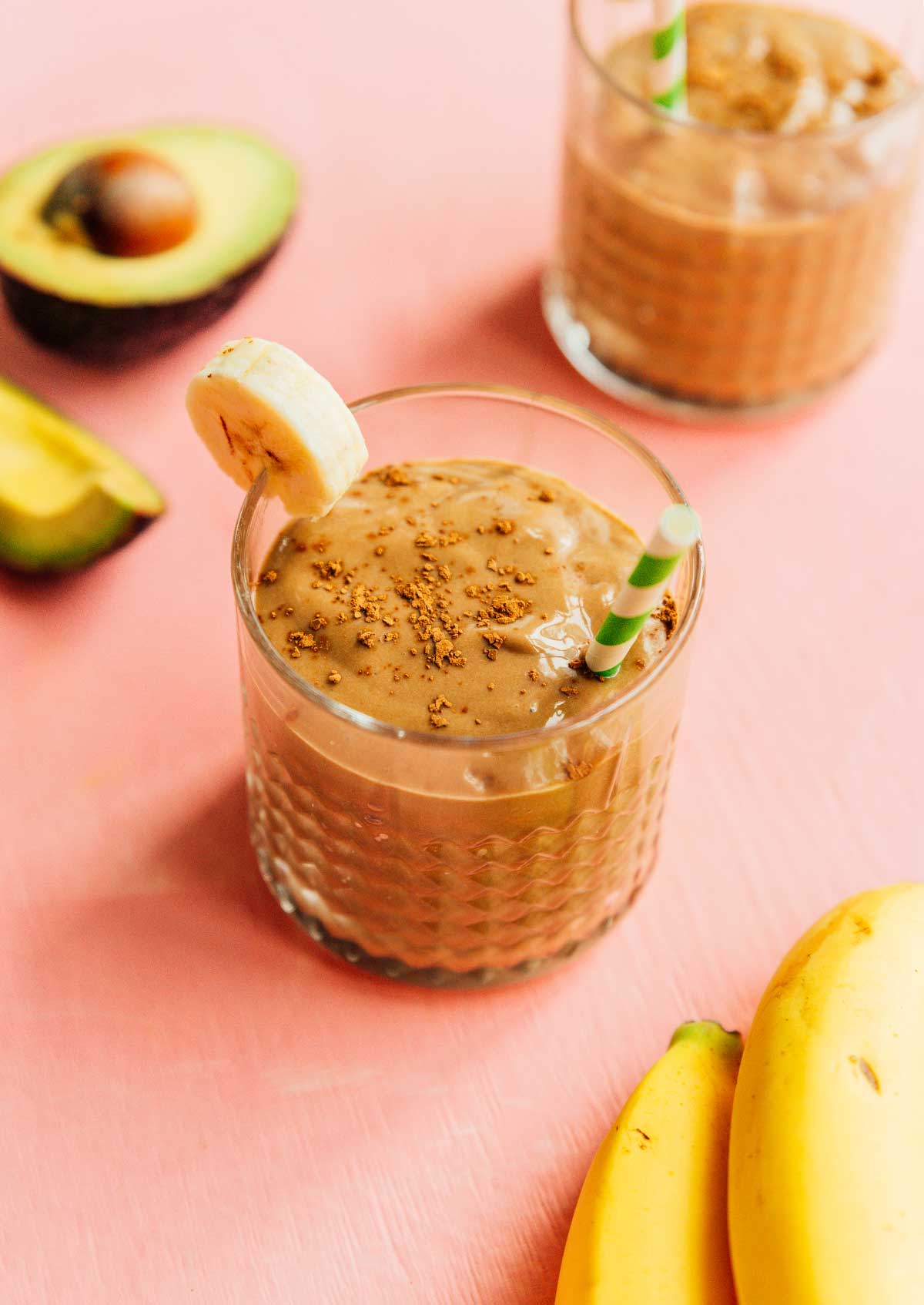 A glass filled with avocado chocolate smoothie and topped with cinnamon, a banana slice, and a green and white striped straw