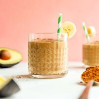 A glass of rich chocolate smoothie garnished with a banana slice and a green straw. Avocado halves hint at the creamy goodness inside, with a spoonful of cocoa powder nearby. Set against a playful pink background, this avocado smoothie is as delightful as it looks.