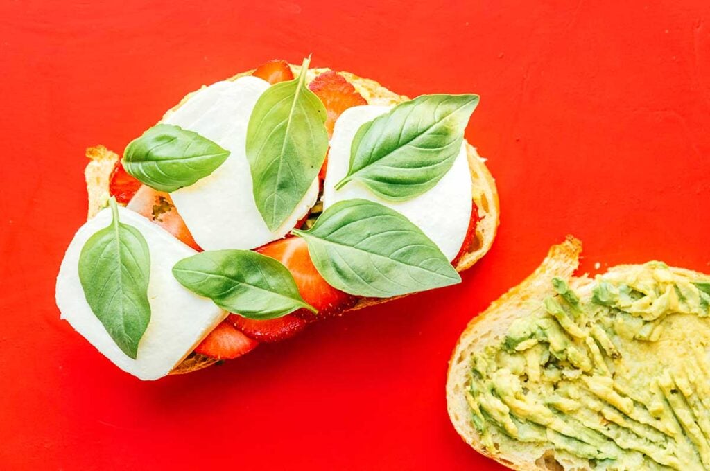 Two slices of avocado bread topped with avocado and one half topped with strawberries, mozzarella, and basil