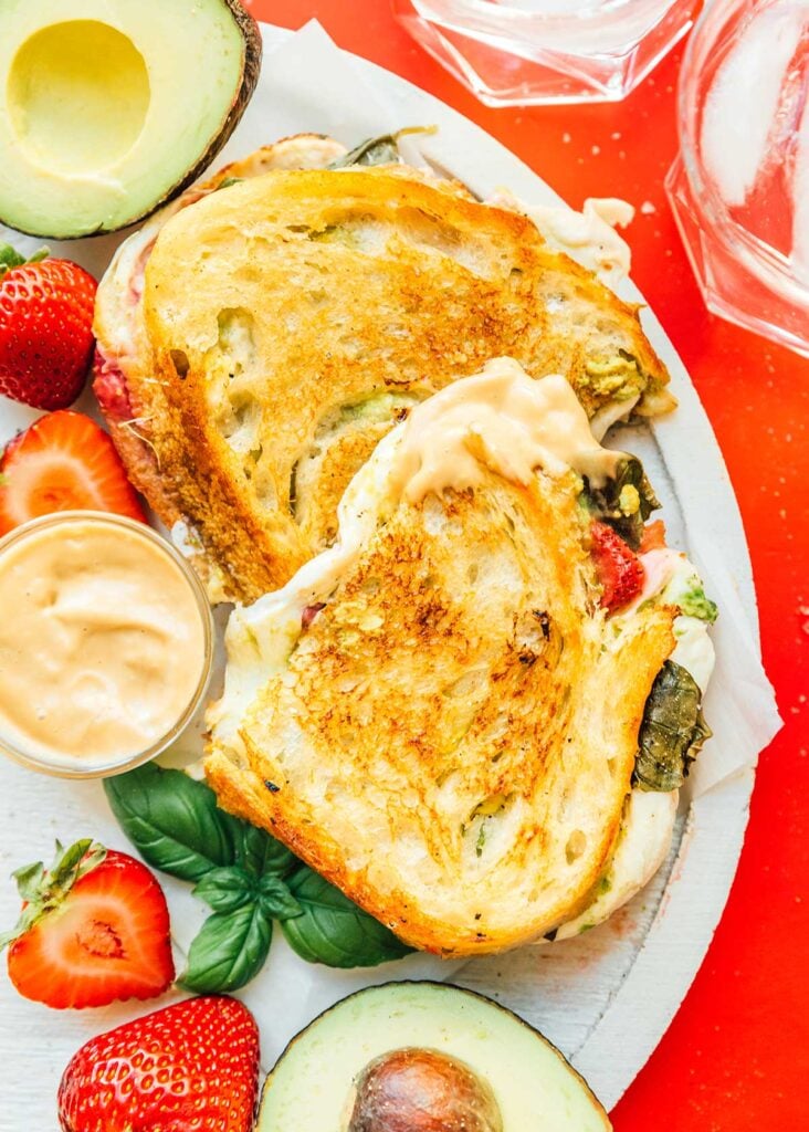 A serving tray filled with a halved strawberry avocado grilled cheese and surrounded by ingredients like strawberries, avocado halves, and a bowl of balsamic mayo dipping sauce