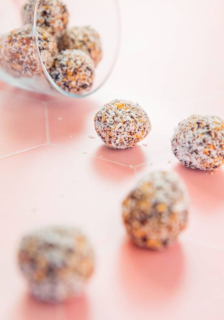 Coconut energy bites scattered on a pink background beside a glass filled with addition bites