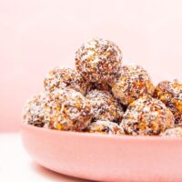 A pink bowl brimming with chocolate no bake energy bites, each delicately coated in shredded coconut, set against a soft pink background.