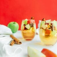 Two glasses of apple sangria with apple cubes and cinnamon sticks sit on a white surface. A green apple, cinnamon sticks, and a white cloth are nearby, set against a vibrant red background.