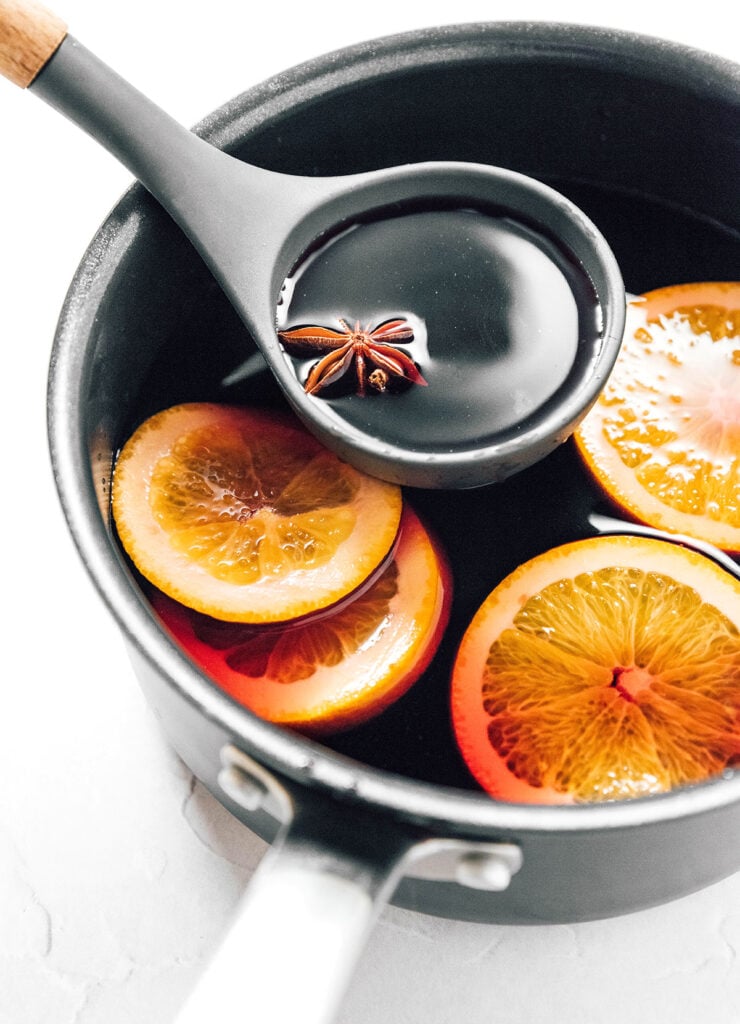A pot filled with steeping gluhwein and orange slices with a ladle scooping out a spoonful that includes a star anise