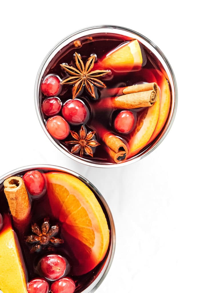 A bird's eye view of two glasses filled with gluhwein mulled wine garnished with cranberries, cinnamon sticks, orange slices, and star anise