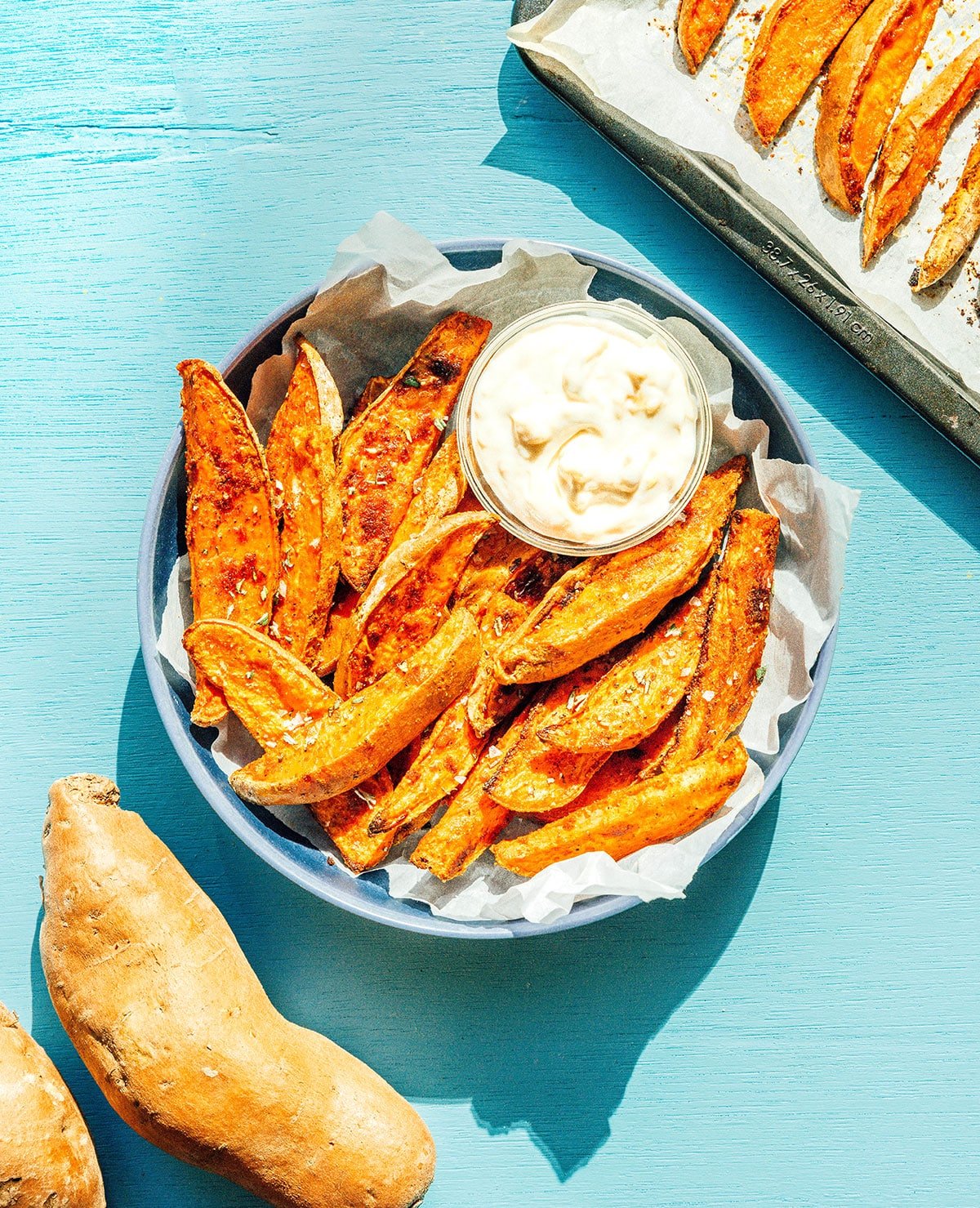 A blue bowl filled with oven-baked sweet potato wedges and a small bowl of garlic mayo sauce