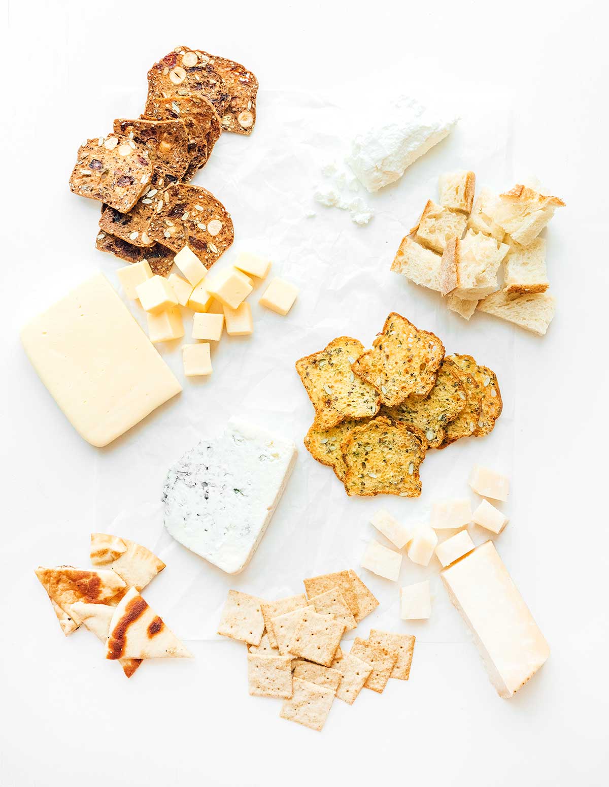 The start to a cheese board with 4 types of crackers and bread pieces spread out in neat groups with four types of cheese placed in between