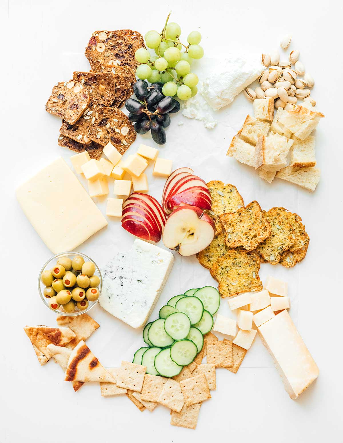 A charcuterie cheese platter that is coming together with crackers, breads, fruits, veggies, cheeses, nuts, and olives