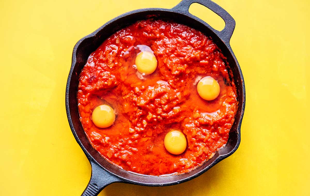Raw eggs in tomato sauce for shakshuka in a cast iron skillet on a yellow background
