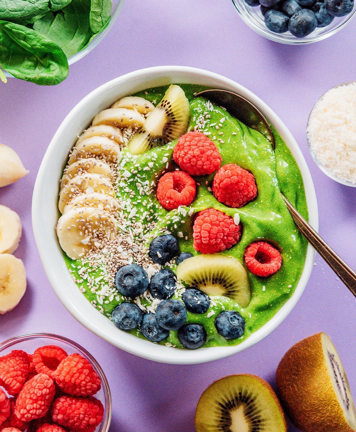 Green smoothie bowl with bananas, kiwi, blueberries, raspberries, coconut, and seeds on top.