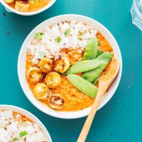 A bowl of red lentil and banana curry is served with rice, snap peas, and grilled vegetables on a teal surface, accompanied by a wooden spoon.