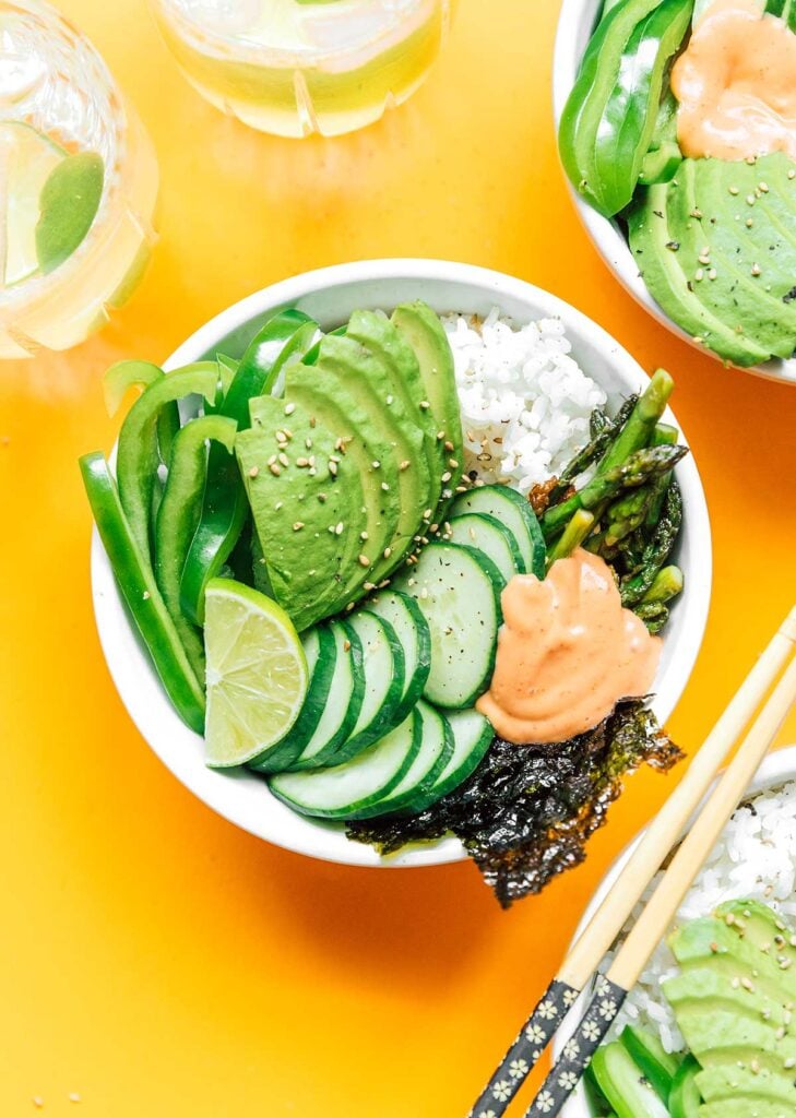 A complete green goddess sushi bowl containing rice, asparagus, sushi paper, cucumber, avocado, green bell pepper, lime, and sriracha mayo