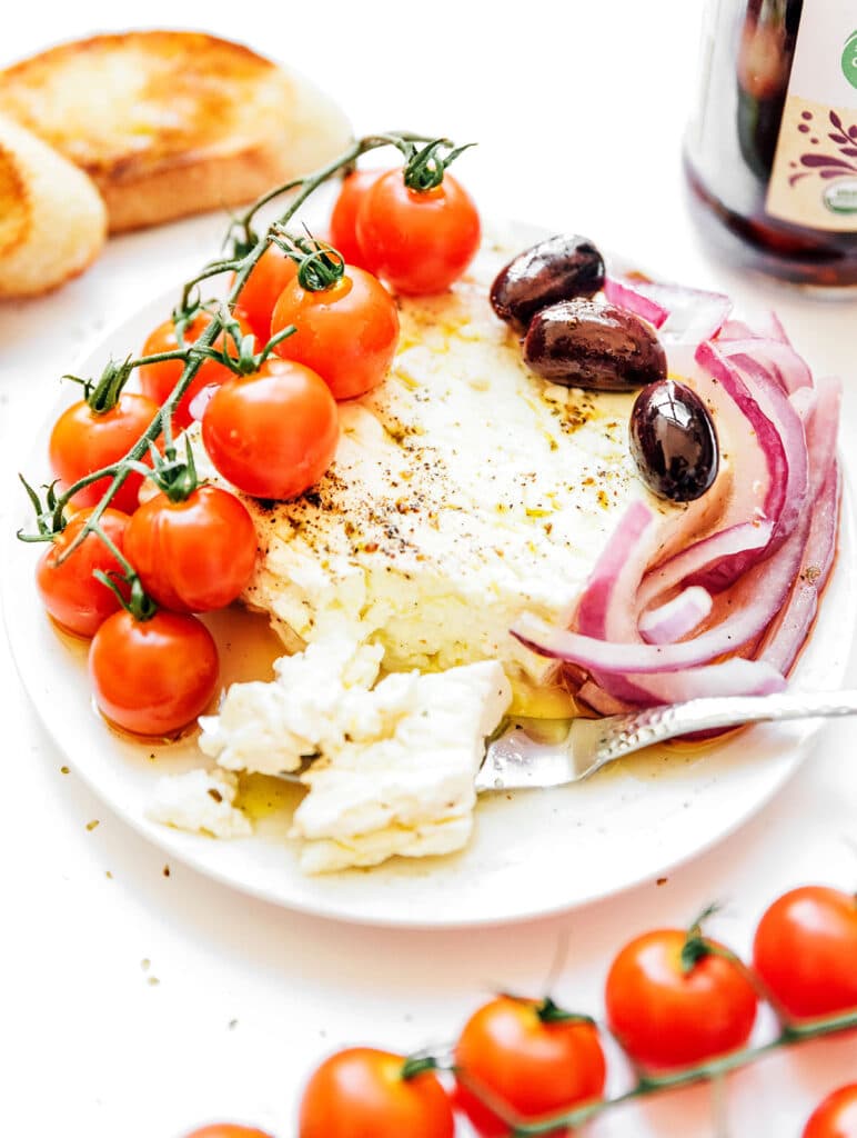 A white plate filled with a wedge of feta cheese, tomatoes, olives, red onion, and a knife cutting off a piece of the feta