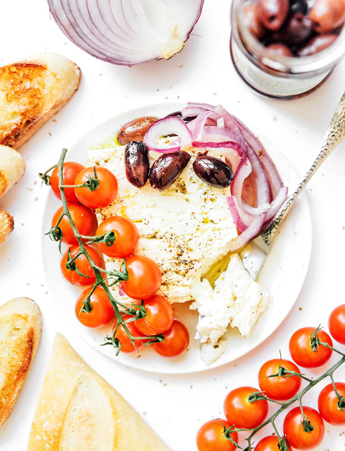 Greek baked feta on a white plate surrounded by ingredients lime tomatoes, red onion slices, and olives.
