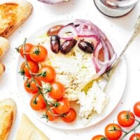 Baked feta on a plate with onions, olives, and tomatoes along side it.