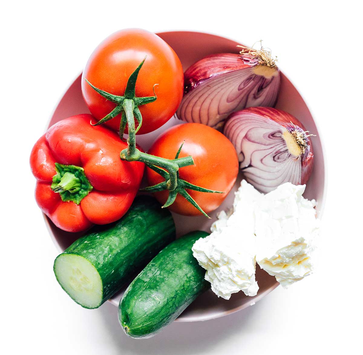 A bowl filled with two tomatoes, a red onion chopped in half, two blocks of feta cheese, a cucumber chopped in half, and a red bell pepper