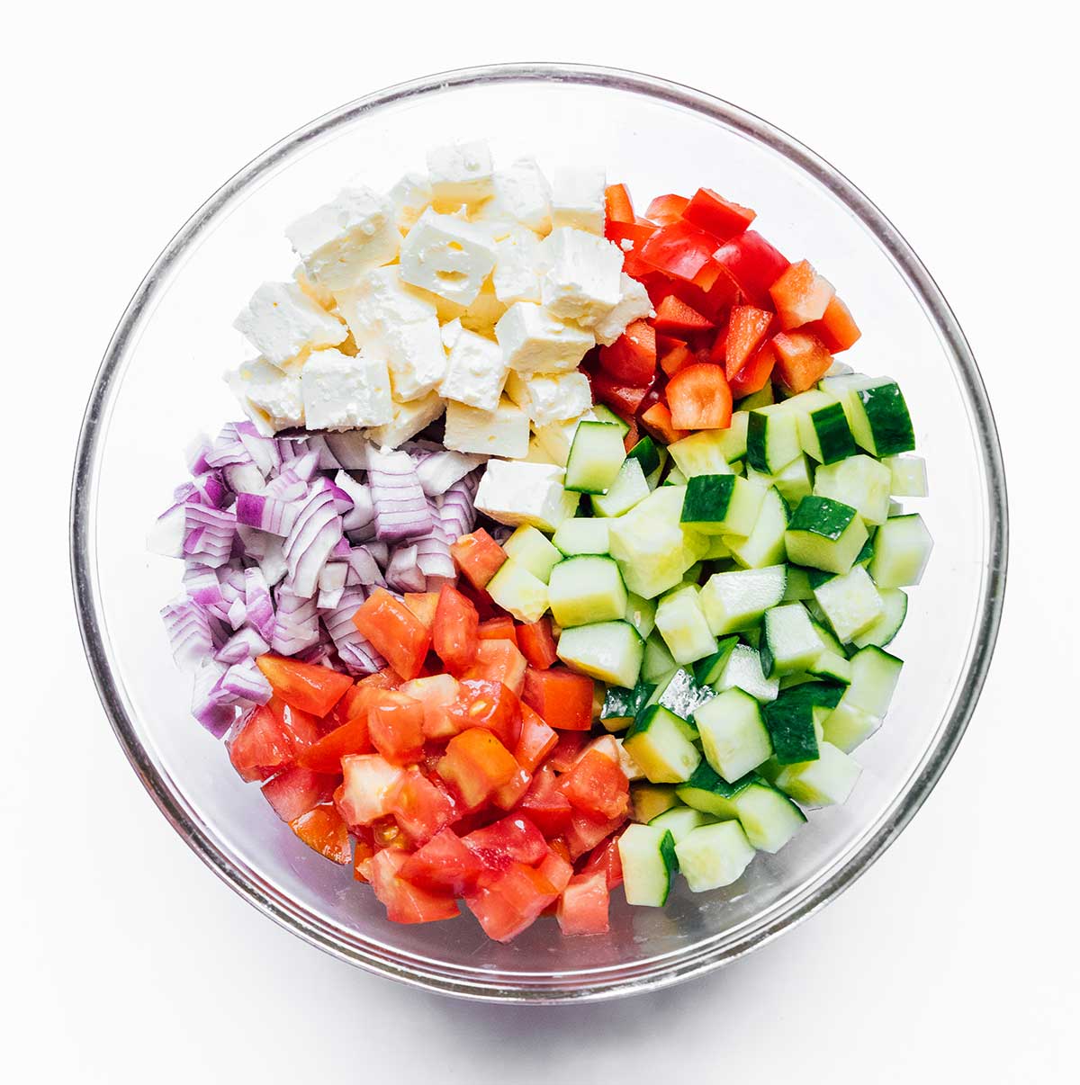 A glass dish filled with chopped salad ingredients including feta, red bell pepper, cucumber, tomatoes, and red onion