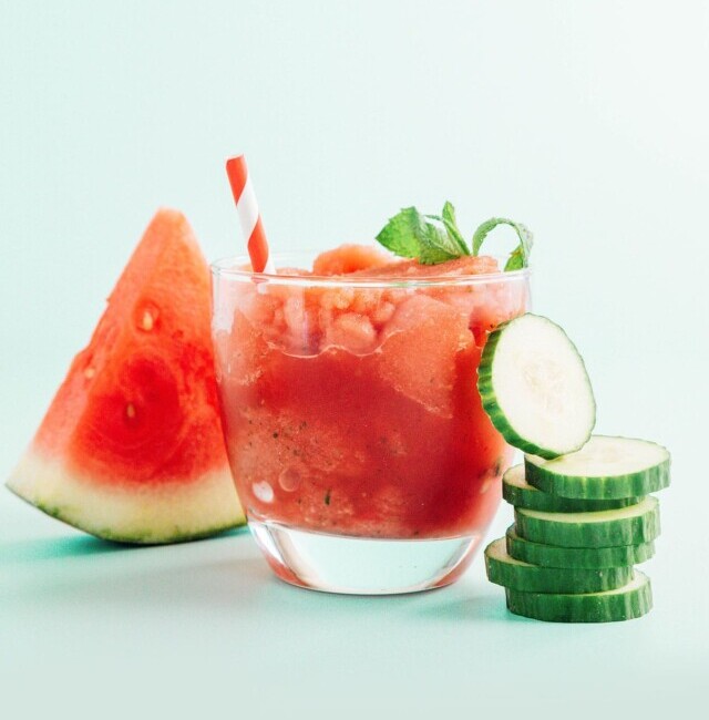A red smoothie in a glass with a red straw and a watermelon slice and stack of cucumbers next to it.