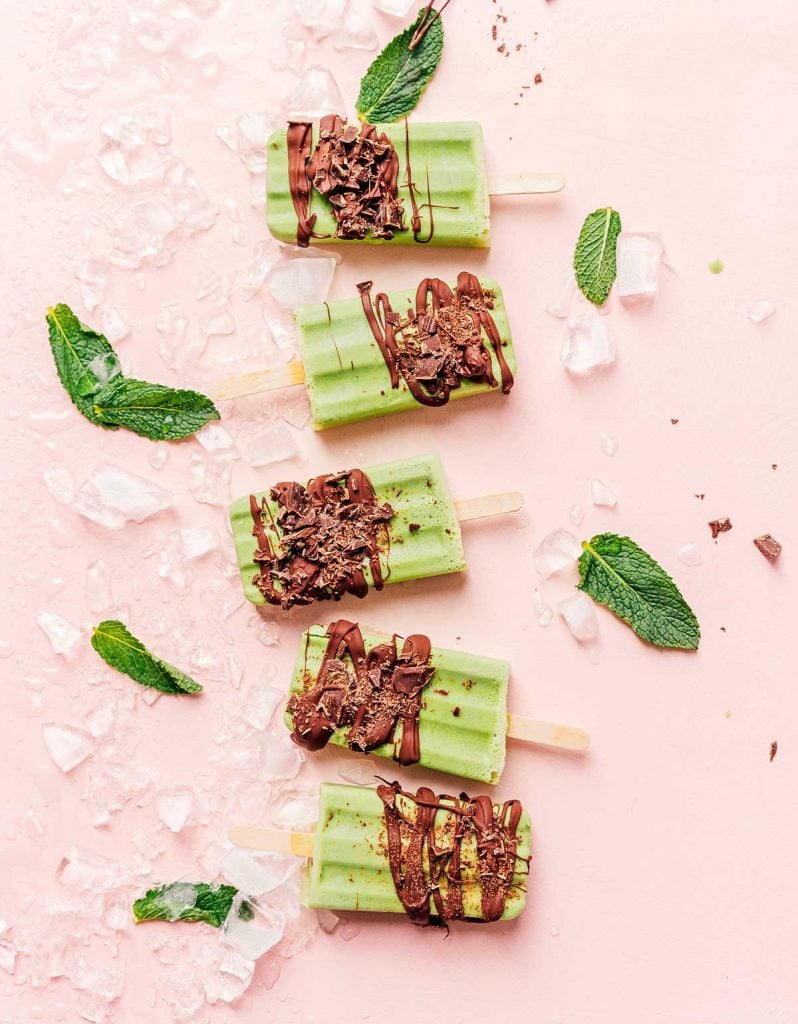Green mint chocolate popsicles on a pink background with ice and mint leaves