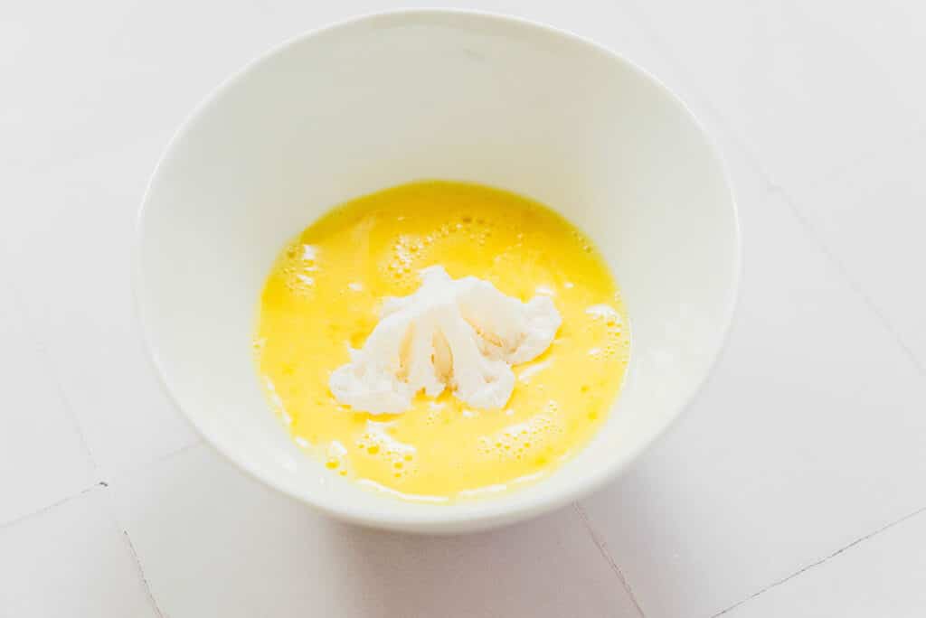 One cauliflower floret being dipped in a white bowl filled with an egg wash