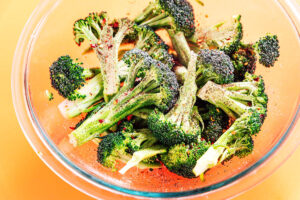 A clear glass bowl filled with roasted broccoli florets seasoned with ground black pepper and red pepper flakes on a vibrant yellow background.
