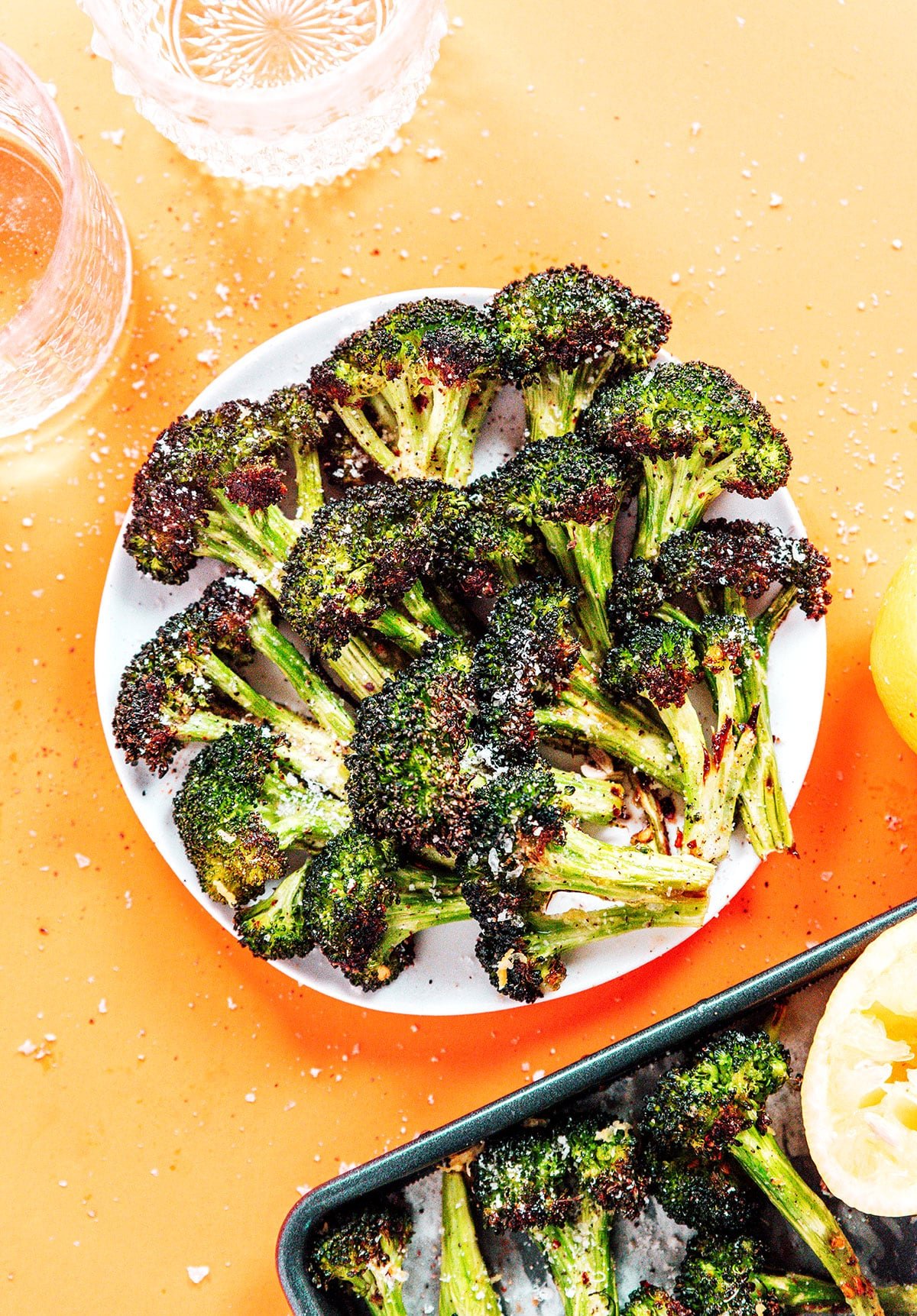 Roasted broccoli on a white plate with salt and spices surrounding them and a sheet pan off to the side.