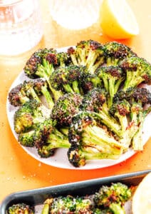 A plate of roasted broccoli florets sprinkled with grated cheese or seasoning, served with a halved lemon and a glass of water nearby.