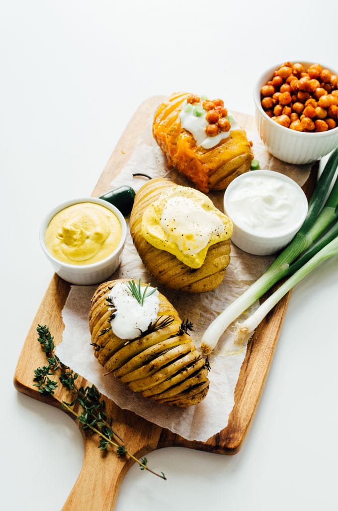 Hasselback potatoes on a cutting board - Change up the routine and make these easy hasselback potatoes for dinner tonight! We're whipping up three tasty flavors: vegan nacho, herb, and loaded.