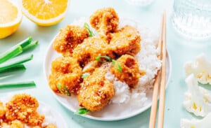 A plate of white rice topped with breaded orange cauliflower, sauced and garnished with sliced green onions; chopsticks, orange halves, and scallions are nearby.