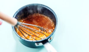 A whisk stirring a thick, orange-brown sauce for orange cauliflower in a small black saucepan, with steam rising from the mixture.