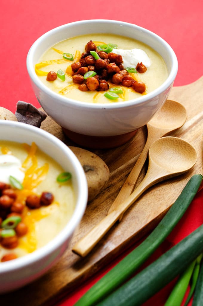 Potato soup in a white bowl on a cutting board on a red background - This Healthy Slow Cooker Potato Soup is a lower calorie, vegetarian alternative (that doesn't compromise on flavor one bit!) Loaded with smoky roasted chickpeas, Greek yogurt, and sharp cheddar.