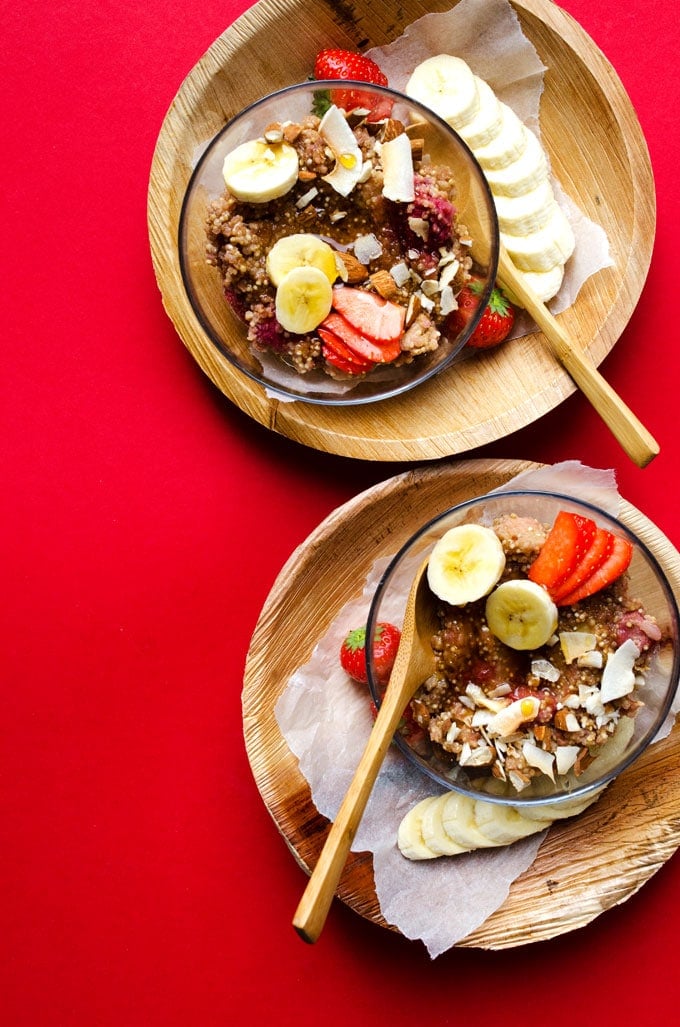 Quinoa in a clear bowl on a red background with wooden spoons - This Overnight Quinoa Breakfast Porridge is a delicious excuse to break out your slow cooker. It tastes like strawberries and cream (while being a healthy, protein-packed way to start your day!)