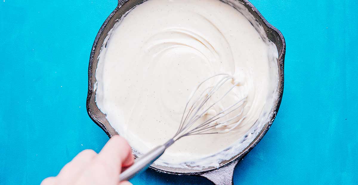 Whisking bechamel sauce in a cast iron skillet with a blue background
