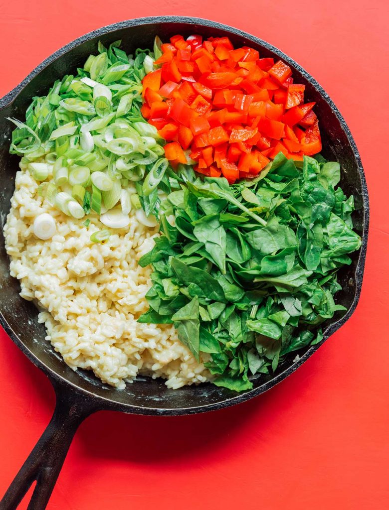 A cast iron skillet filled with Arborio rice, green onion, red bell pepper, and spinach