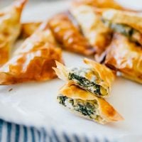 How to make spanakopita, cut opne on a white plate with a blue towel - These Spanakopita Triangles are a simple appetizer jam-packed with feta and spinach, wrapped up in buttery, flaky phyllo crust.