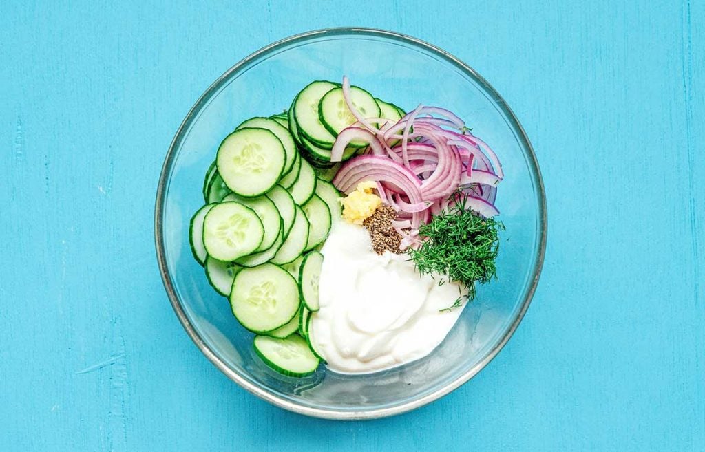 A glass bowl filled with creamy cucumber salad ingredients including cucumber, red onion, fresh dill, Greek yogurt, and garlic