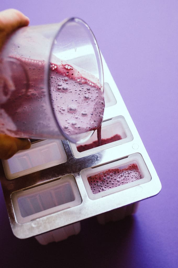 Pouring popsicle mixture into mold on purple background - These Blueberry Cheesecake Popsicles are a creamy and refreshing summer snack that are low in calories, ingredients, and prep time (but certainly not low on that classic blueberry cheesecake flavor!) Dig out your popsicle mold and give these a go.