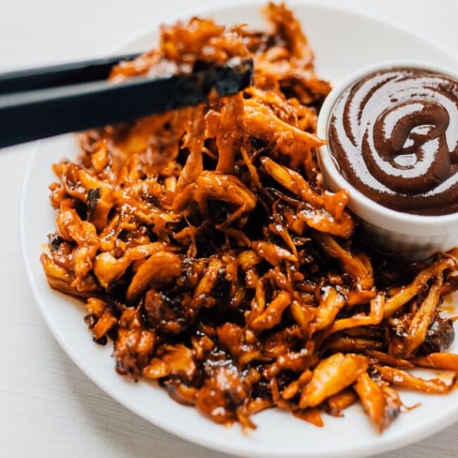 Vegan mushroom pulled pork on a white background and white plate - By shredding king oyster mushrooms, seasoning with spices, and baking, you can create a vegan mushroom pulled pork that rivals the real stuff! Perfect on sandwiches, tacos, nachos...or whenever you need pulled pork.