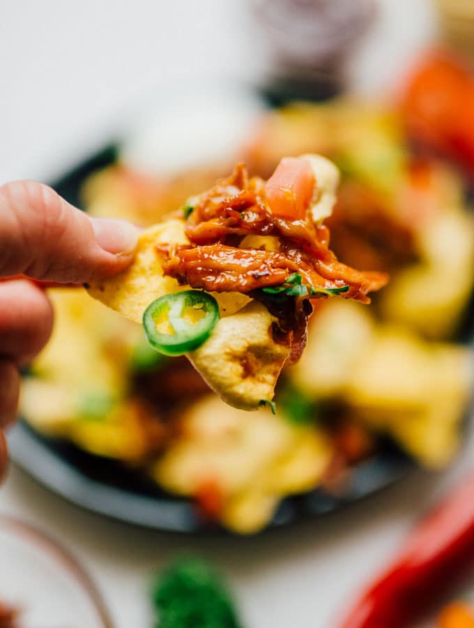 Closeup of nacho chip in hand - These vegan nachos are piled high with easy mushroom BBQ "pulled pork" and a cashew-based queso cheese sauce that will knock your dairy-free socks off.